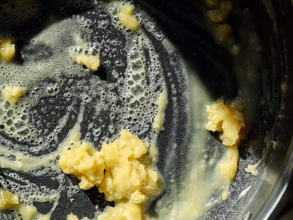 Melted butter and flour mixture sizzling in a pan, forming a light roux with some lumps. Bubbles appear around the edges as the mixture cooks, creating a swirl pattern in the pan.